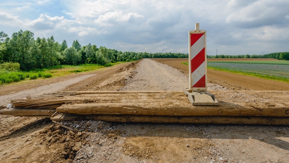 Road under construction closed with a wooden beam and a red and white road sign. The road is located on top of newly built Dutch dike. In the background is a field of cabbage cultivation.